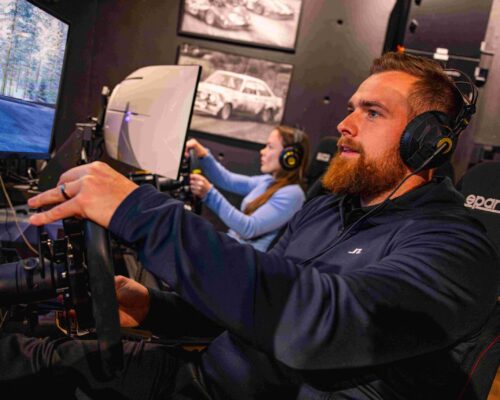 10 Parrakas mies kuvan etualalla ja nainen sivulla ajavat kilpaa ajosimulaattoreilla.