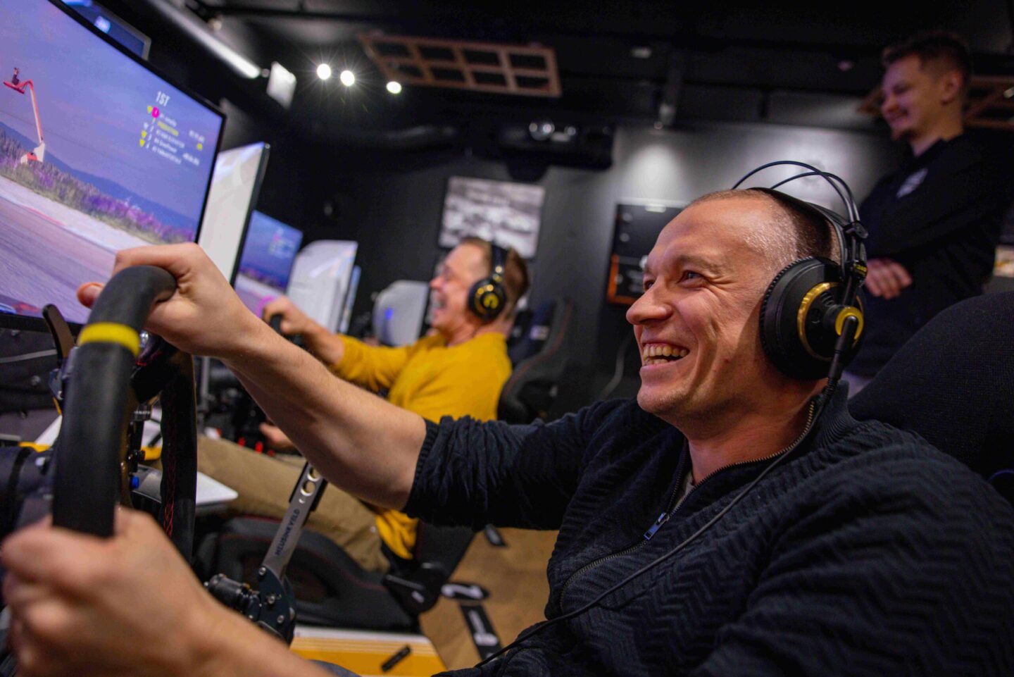 Kaksi miestä pelaa rallisimulaattoria rinnakkain Shakedown Parkissa iloisina, taustalla kolmas henkilö seuraa hymyillen tilannetta.